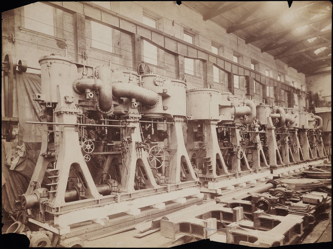En svartvit vintagefotografi som visar en rad stora industrimaskiner, troligen ångmaskiner eller tillverkningsutrustning från tidigt 1900-tal, uppställda i en fabriksmiljö med stora fönster och industriell arkitektur.